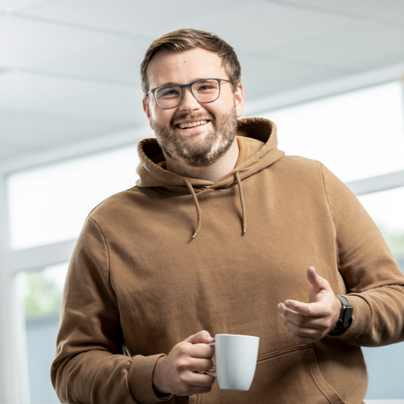 Optitime Mitarbeiter Julian Oellers lachend mit einem Kaffee in der Hand